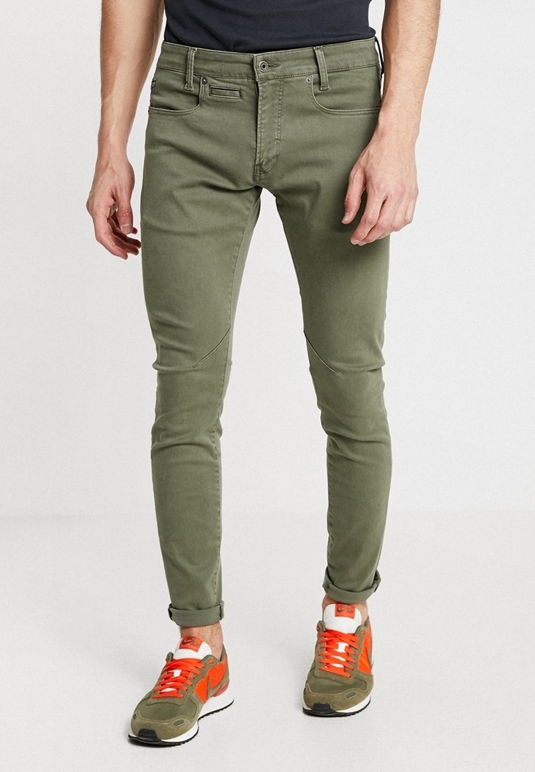 Man som bär slim-fit olivgröna byxor och matchande olivgröna och orange sneakers, stående mot en enkel bakgrund.