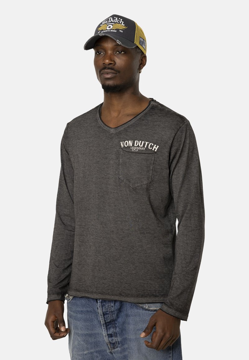 Chemise à manches longues gris foncé avec un col en V et une poche poitrine ; présente le logo blanc "Von Dutch". Associée à un jean délavé bleu clair et une casquette.