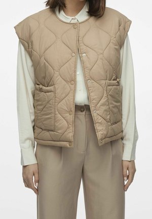 Kvinde iført beige quiltet ærmeløs vest over en hvid skjorte med knapper og beige bukser med vidde i benene, stående med afslappede arme.