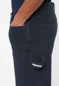 Pánska ruka v prednom vrecku námorníckych modrých textúrovaných nohavíc s extra bočným vreckom, ktoré má červenobielo-modrý prúžkovaný popruh.