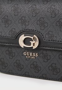 Fekete textúrájú kézitáska arany színű G logós csattal és "GUESS EST1981" felirattal, fényesen emboszálva az elülső részén.