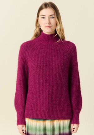 Fuksia neulepusero korkealla kauluksella, teksturoidulla kuviolla ja pitkillä hihoilla. Ribbaatut helma ja hihansuut. Asetettu päälle monivärisen gradient-hameen.