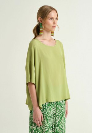 Femme en haut ample vert clair et jupe verte à motifs, portant de longues boucles d’oreilles multicolores en perles, debout devant un fond uni.