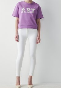 Lila croppad sweatshirt med "ART" i vit text, parat med slim fit vita jeans och beige spetsiga skor. Släta texturer genom hela outfiten.