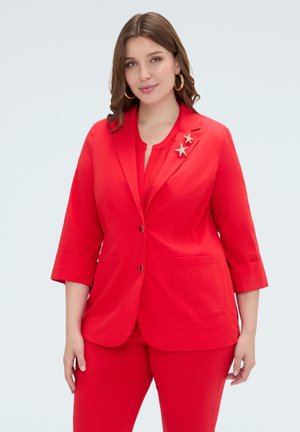 Donna con capelli castani fino alle spalle indossa un abito rosso brillante con spille a forma di stella marina sulla rever, su uno sfondo chiaro.