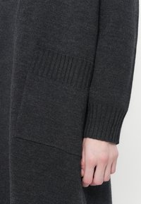 Cardigan gris en tricot avec des poignets côtelés et des poches, affichant une texture douce avec un motif subtil intégré dans le tissu.