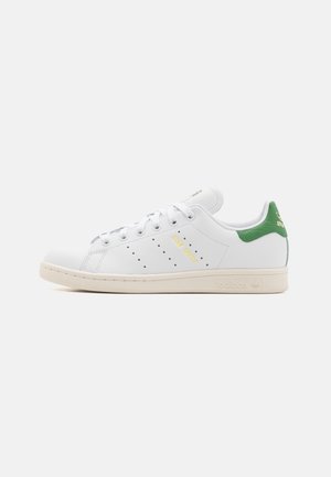Zapatilla Adidas Stan Smith blanca con talón verde, franjas perforadas y suela beige, mostrada desde el lateral sobre un fondo blanco.