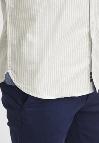 Lys farget stripete skjorte med avrundet kant, laget av mykt stoff. Mørk marineblå bukser med subtil tekstur og synlig sidelomme.