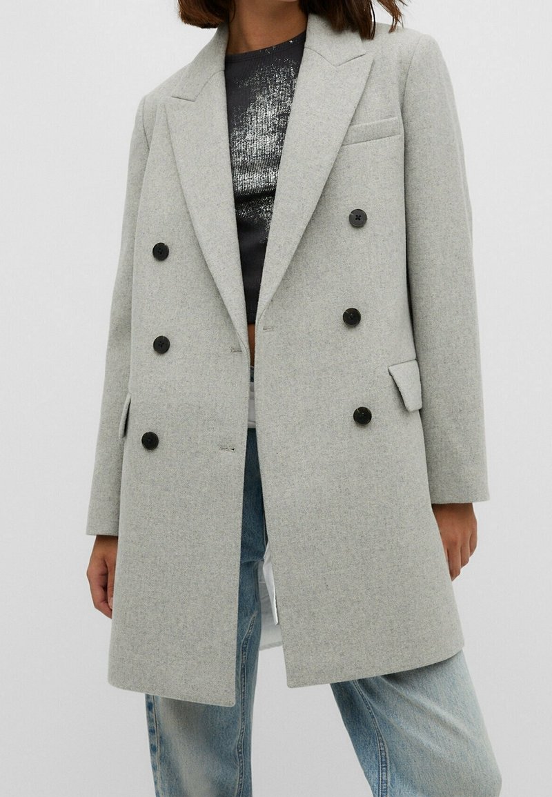 Mujer vestida con un abrigo gris claro de doble botonadura sobre una blusa negra y plateada, con jeans de color azul claro, frente a un fondo sencillo.