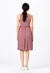 Šaty bez rukávů v jemné levandulové barvě, pletený trup s volánkovým páskem, splývavá látka níže, délka po kotníky, kombinováno s bílými botami.