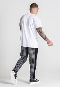 Wei�ßes Baumwoll-T-Shirt kombiniert mit schwarzen gemusterten Jogginghosen mit geometrischem Design und seitlichen Streifen. Die Schuhe sind schwarz mit weißen Sohlen.