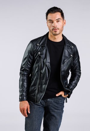 Giacca da motociclista in pelle nera con zip asimmetrica, ferramenta argentata e design aderente, abbinata a una camicia nera e jeans blu.