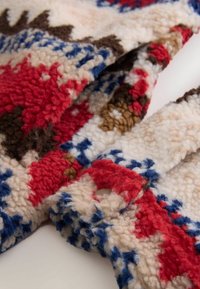 Uldent stof med et mønster i rød, blå, brun og creme. Tekstureret overflade med bløde, fluffy fibre og en knapdetalje.