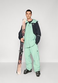 Abbigliamento da sci con giacca e pantaloni verde menta, e cappuccio e dettagli blu navy. Uno sciatore tiene in una mano sci neri con attacchi.