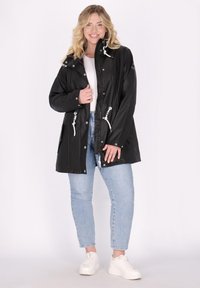 Giacca a vento nera con cappuccio regolabile, vita con cordino e due tasche frontali. Abbinata a jeans azzurri e scarpe da ginnastica bianche.