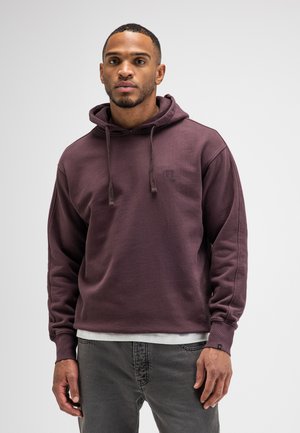 Mann trägt kastanienbraunen Kapuzenpullover und graue Jeans, steht vor einem einfarbigen hellen Hintergrund.