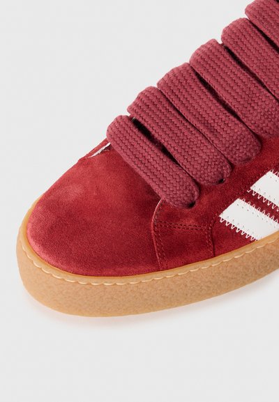 Red suede sneaker with thick braided laces, white stripes, and a textured gum sole. The toe is rounded, and the stitching is visible.
