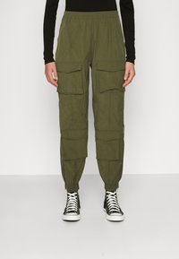 Olivgröna cargo-byxor med flera framfickor, avsmalnande anklar, elastisk midja och dragkedjor vid bensluten, tillsammans med svarta sneakers.