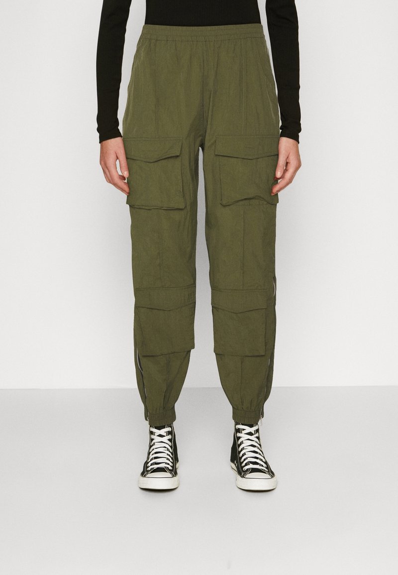 Olivgröna cargo-byxor med flera framfickor, avsmalnande anklar, elastisk midja och dragkedjor vid bensluten, tillsammans med svarta sneakers.