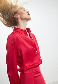 Camicia rossa in satin a maniche lunghe con colletto, con dettagli di bottoni e una silhouette elegante. Sfondo bianco e semplice.
