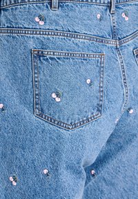 Šviesiai mėlyni džinsai su rožinėmis siuvinėtomis vyšniomis ir žaliais stiebais, su kišene ant nugaros. Klasikinis penkių kišenių dizainas.