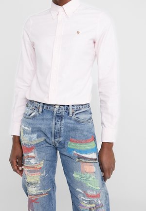 Camisa abotonada de rayas rosa claro con cuello, combinada con jeans azules desgastados con parches de colores y reparaciones visibles.