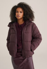 Plūmkrāsas pūkainais jaka ar kapuci, ar quilted dizainu, priekšējo rāvējslēdzēju un sānu kabatām. Valkāts virs struktūrēta vīna sarkanā džempera.