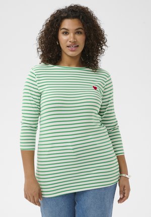 Woman with curly hair wearing a white and green striped shirt with a small red heart on the chest, standing against a white background.