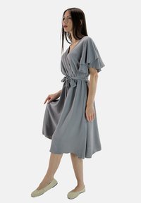 Robe grise longueur genou avec un design croisé, manches courtes à volants, et une ceinture ajustée à la taille. Accompagnée de ballerines de couleur claire.