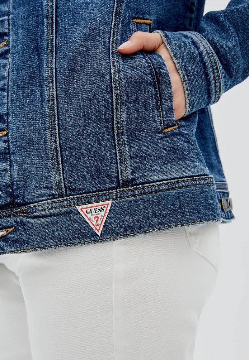 Giacca di denim blu scuro con un patch triangolare del logo "Guess", cuciture in contrasto, tasche e una vestibilità su misura, abbinata a pantaloni bianchi.