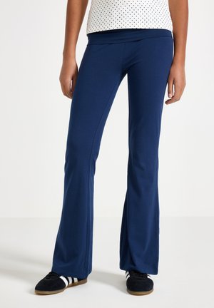 Nedre del af en person iført marineblå udvidede bukser, hvidt prikket top og sorte sneakers med hvide striber på hvid baggrund.