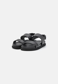 Sandali in pelle nera con ampie cinghie incrociate, fibbia alla caviglia regolabile, suola piatta e soletta texturizzata per il massimo comfort.