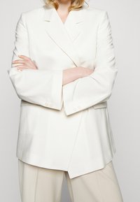 Biała dwurzędowa marynarka z szerokimi klapami, długimi rękawami i zapinanymi na guziki mankietami. Gładka faktura i luźny krój, zestawiona z neutralnymi spodniami.