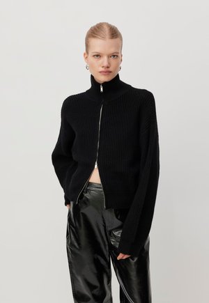 Young woman wearing a black ribbed zip-up sweater and shiny black leather pants against a plain light background.