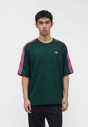 Ung mand iført mørkegrøn Adidas T-shirt med pink striber på ærmerne og pink logo, stående foran en ensfarvet hvid baggrund.