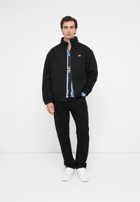 Μαύρο bomber μπουφάν με ψηλό γιακά, ραμμένες μανσέτες και μικρό λογότυπο. Φορεμένο πάνω από μπλούζα με μαύρες και μπλε ρίγες, σε συνδυασμό με μαύρο παντελόνι και λευκά παπούτσια.