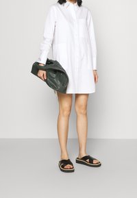 Donna che indossa un vestito camicia bianco con bottoni, tiene in mano una pochette di pelle verde scuro e indossa sandali con zeppa neri.