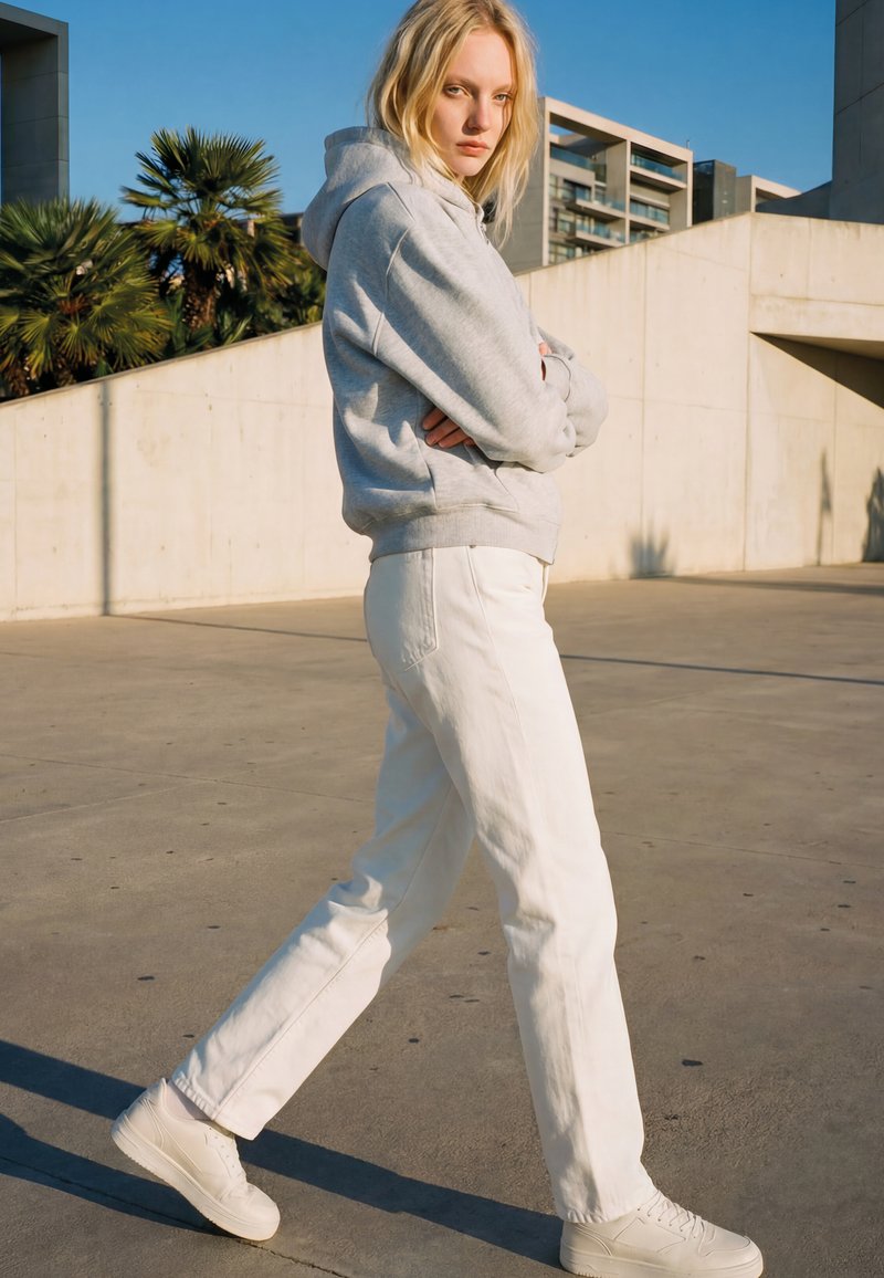 Femme blonde portant un sweat à capuche gris, un pantalon blanc et des baskets blanches marchant en plein air près de bâtiments modernes en béton sous un ciel dégagé.