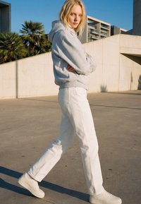 Femme blonde portant un sweat à capuche gris, un pantalon blanc et des baskets blanches marchant en plein air près de bâtiments modernes en béton sous un ciel dégagé.