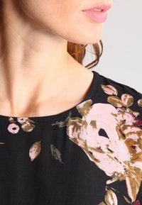 Bluse mit floralem Druck, runder Ausschnitt, dunkler Hintergrund, große Rosenmuster in Rosa-, Gold- und Grüntönen. Weicher Stoff.