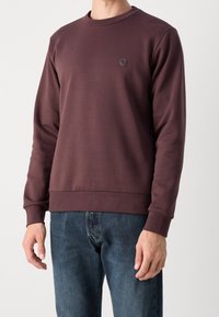 Sudadera de color burdeos con cuello redondo, puños y dobladillo acanalados. Presenta un pequeño parche negro con el logo en el lado izquierdo del pecho. Combinada con vaqueros azules.