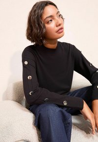 Pulover negru din tricot cu guler cu nervuri și detalii cu nasturi pe mâneci. Purtați-l cu pantaloni din denim albastri, așezat pe o suprafață texturată de culoare deschisă.