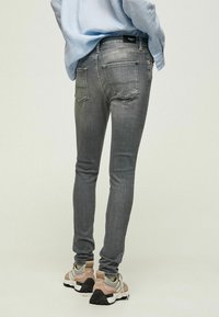 Jeans de mezclilla gris ajustados con una textura desgastada, cinco bolsillos y un logotipo bordado en la cinturilla, combinados con zapatillas de colores claros.