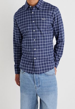 Man draagt een blauw-wit geruit overhemd met knopen en lange mouwen, en lichtblauwe spijkerbroek, staat met één hand in de zak.