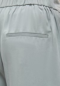 Κοντινή λήψη ανοιχτογκρι παντελονιού με ελαστική μέση και μία οριζόντια πίσω τσέπη με διακριτική ραφή.