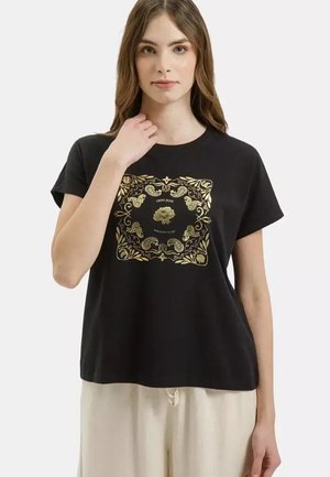 Młoda kobieta ubrana w czarny T-shirt ze złotym kwiatowym i paisleyowym wzorem na kwadratowym tle oraz beżowe spodnie, ręka przy dekolcie.