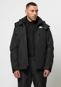 Schwarze wasserdichte Jacke mit Reißverschluss, verstellbarer Kapuze und Reißverschlusstaschen. Verfügt über ein Futter aus Fleece und ein weißes Logo-Detail auf der Brust.