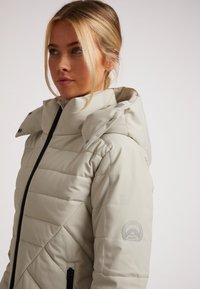 Beige pufferjakke med lynlås og trykknaplukning, quiltet tekstur, justerbar hætte og subtil logo detalje på ærmet.