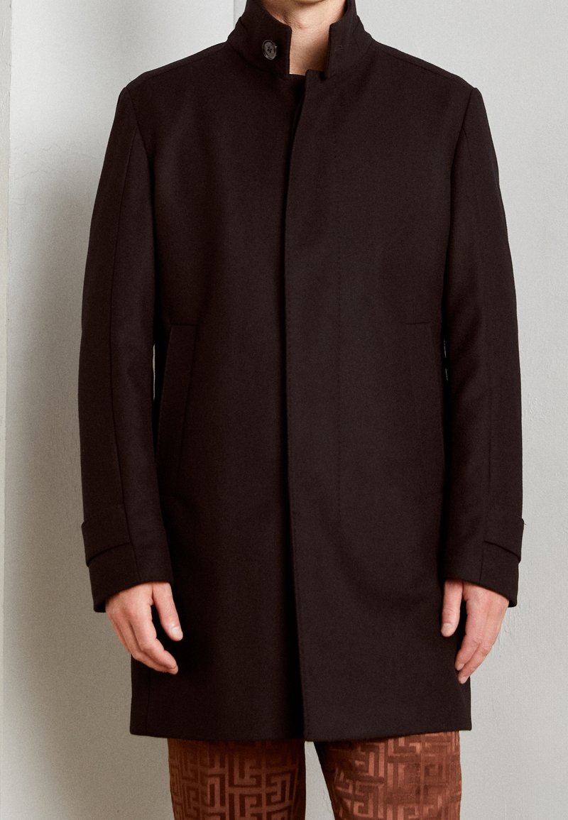 Homme portant un manteau à col montant marron foncé avec fermeture à boutons et un pantalon marron à motifs, sur un fond uni.