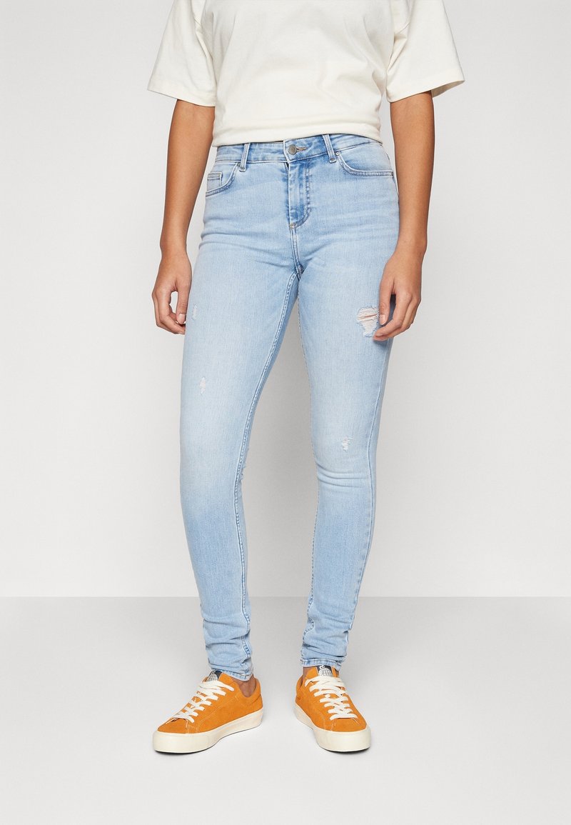 Persona vestida con vaqueros ajustados rasgados de color azul claro, camiseta blanca y zapatillas naranjas, apoyada contra un fondo liso.